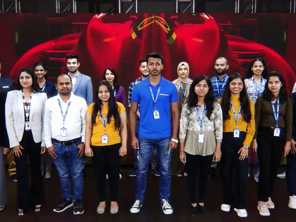 Diverse professional team of achievers in formal and semi-formal attire posing confidently together on stage, illuminated by dramatic red LED backdrop with flowing fabric effects and golden accents, created by Spark VFX Studios for Achievers League corporate event visuals.